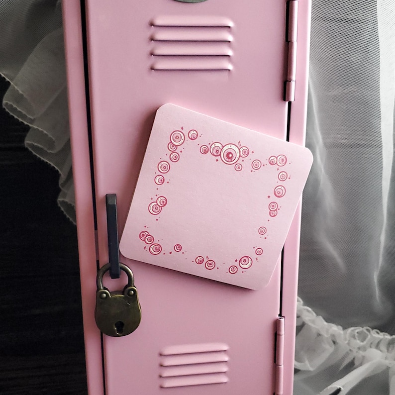 May include: A pink locker with a square pink magnet featuring a decorative design of pink circles. A small, antique-style padlock hangs from the locker door. The locker is a light pink color.