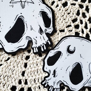 Op de afbeelding: Twee witte stoffen schedelpatches met zwarte borduursels. De ene schedel heeft een pentagram op het voorhoofd en de andere een halve maan.