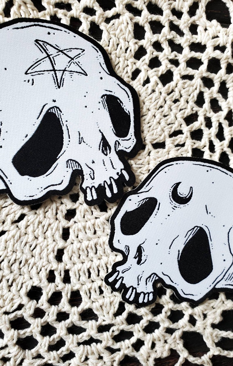 Op de afbeelding: Twee witte stoffen schedelpatches met zwarte borduursels. De ene schedel heeft een pentagram op het voorhoofd en de andere een halve maan.