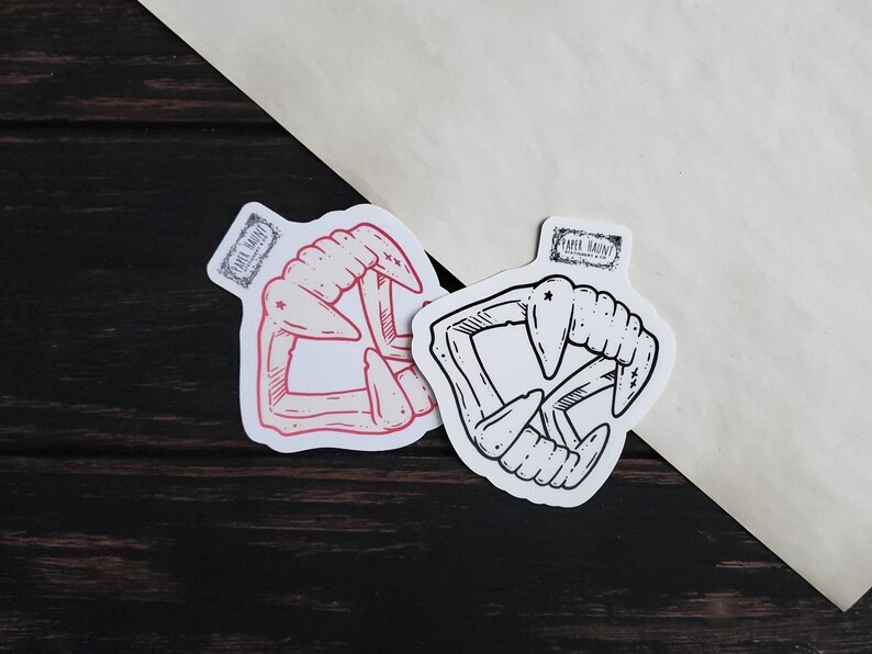 May include: Two sticker designs of a set of fangs, one in black and white and the other in red and white. The stickers are on a white background.