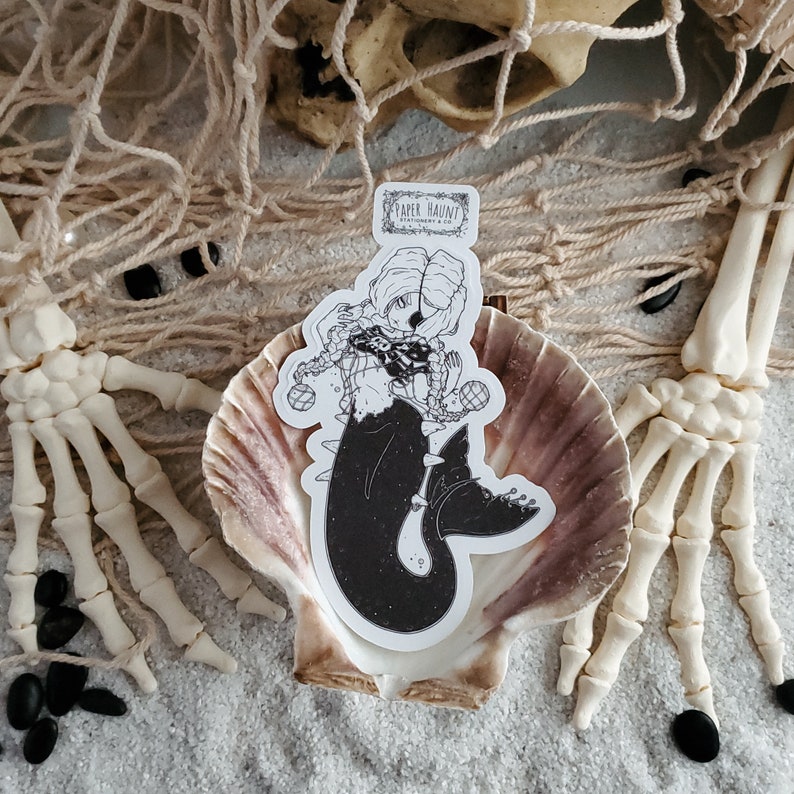 Gothic pirate mermaid sticker by artist White stag for Paper Haunt Stationery & Co in a sea shell on sand with a skull and skeleton hands reaching through a fish net.