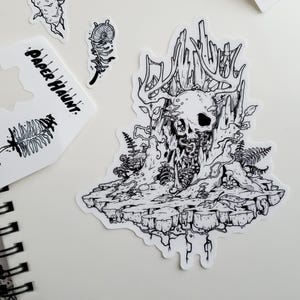 May include: Black and white line art illustration of a skull with antlers, surrounded by vines and mushrooms. The skull is sitting on a rock formation with roots and vines hanging down.