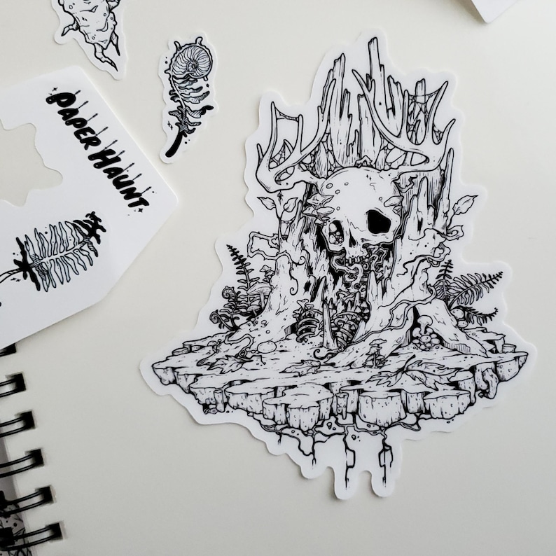 May include: Black and white line art illustration of a skull with antlers, surrounded by vines and mushrooms. The skull is sitting on a rock formation with roots and vines hanging down.