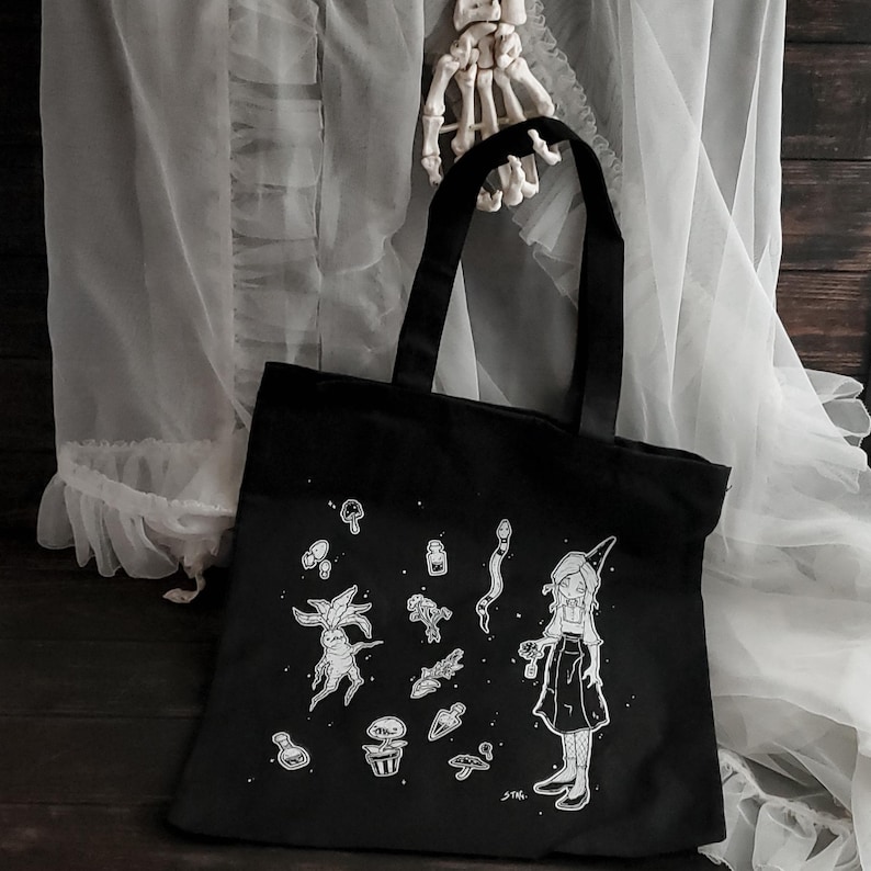 K&ouml;nnte beinhalten: Schwarze Stofftasche mit einer wei&szlig;en Illustration einer Hexe, die eine Zaubertrankflasche h&auml;lt, umgeben von Pilzen, Pflanzen und anderen magischen Gegenst&auml;nden. Der Text "STK." befindet sich in der rechten unteren Ecke.