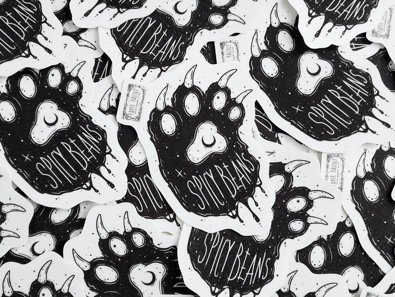 Puede incluir: Una colecci&oacute;n de pegatinas blancas con un dise&ntilde;o de huella de pata negra, con el texto "SPICY BEANS" en una fuente estilizada. La huella de la pata incluye detalles de luna creciente y estrellas. Las pegatinas tienen una est&eacute;tica &uacute;nica, dibujada a mano.