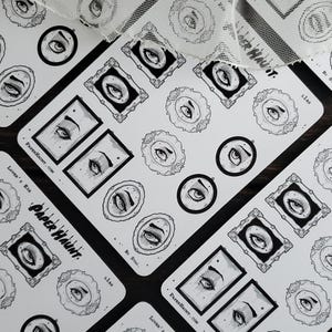 May include: A sheet of stickers featuring black and white illustrations of eyes in ornate frames. The stickers are on a white background and have the text "Paper Haunt" and "Lover's Eye" printed on them.