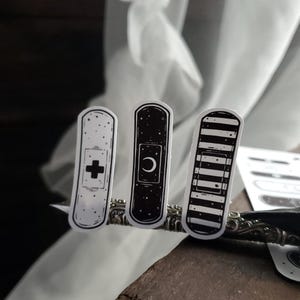 May include: Three black and white band-aid stickers with different designs. The first sticker has a white cross on a black background. The second sticker has a white crescent moon on a black background. The third sticker has black and white stripes.