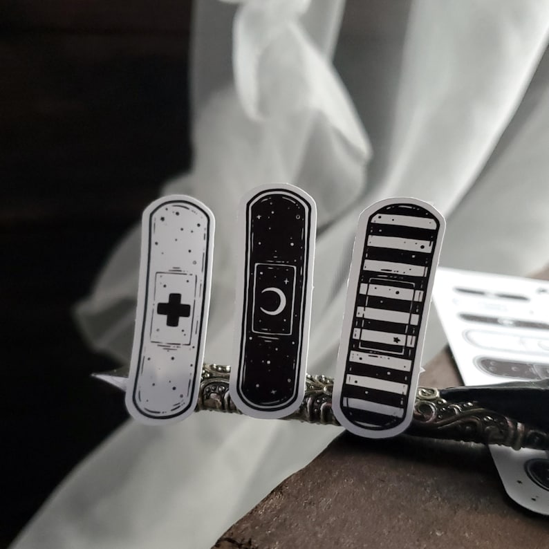 May include: Three black and white band-aid stickers with different designs. The first sticker has a white cross on a black background. The second sticker has a white crescent moon on a black background. The third sticker has black and white stripes.
