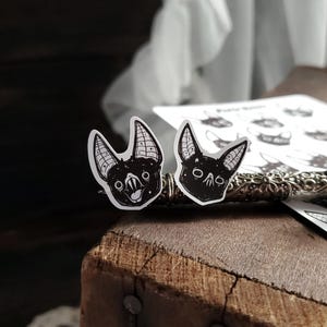 May include: Two black and white bat stickers with a starry background. The bats have different expressions, one is smiling and the other is frowning.