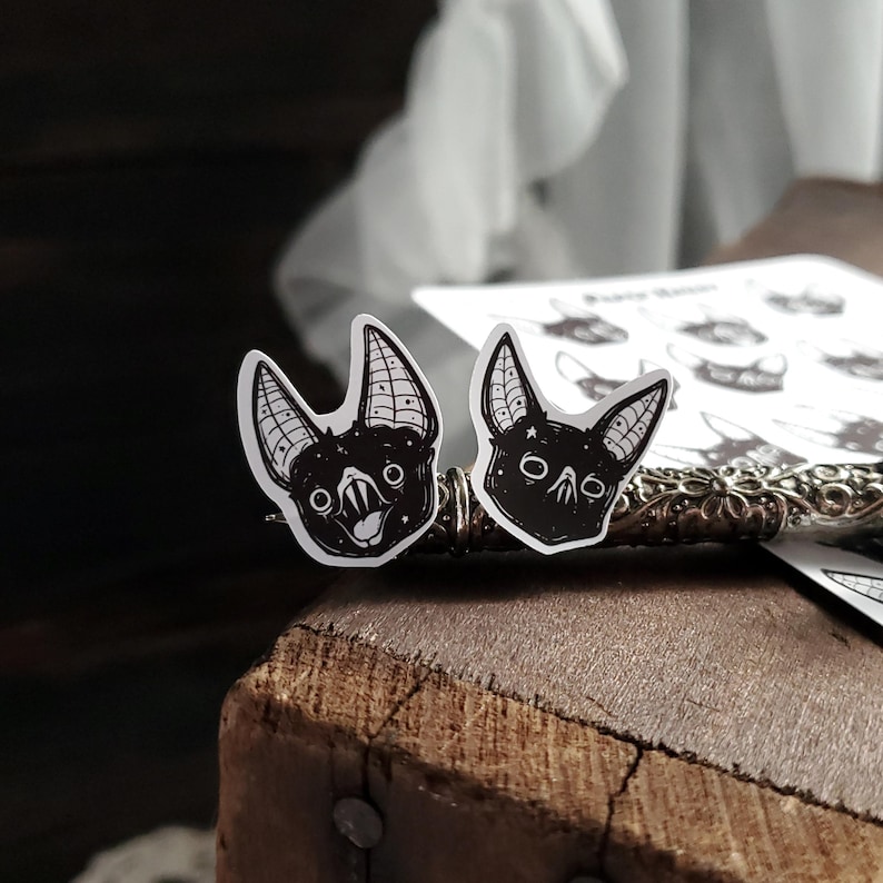 May include: Two black and white bat stickers with a starry background. The bats have different expressions, one is smiling and the other is frowning.