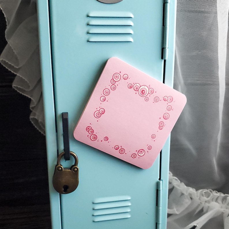 May include: A pink square magnet with a decorative border of circles and dots is attached to a light blue locker. A vintage-style padlock hangs from the locker door. The magnet is a decorative item.