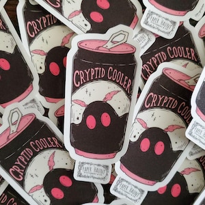 May include: A close-up of several stickers featuring a black and pink can with the text "Cryptid Cooler" and a pink and black creature with two eyes. The stickers are on a white background.