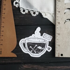 Op de afbeelding: Een zwart-witte sticker van een theekopje met een theezakje waarop "PAPER HAUNT" staat. Het theekopje is gevuld met een donkere vloeistof en er drijven botten in.