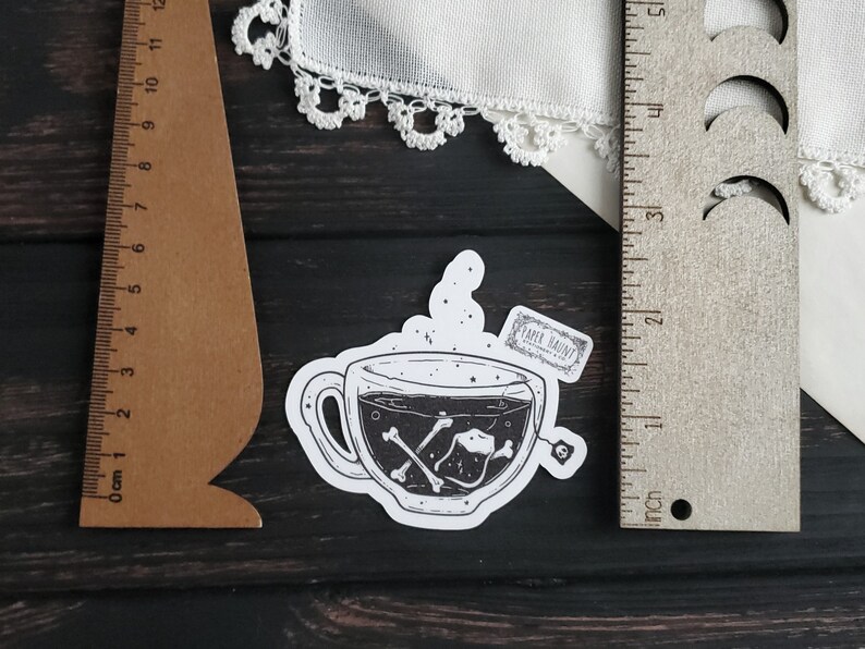 Op de afbeelding: Een zwart-witte sticker van een theekopje met een theezakje waarop "PAPER HAUNT" staat. Het theekopje is gevuld met een donkere vloeistof en er drijven botten in.