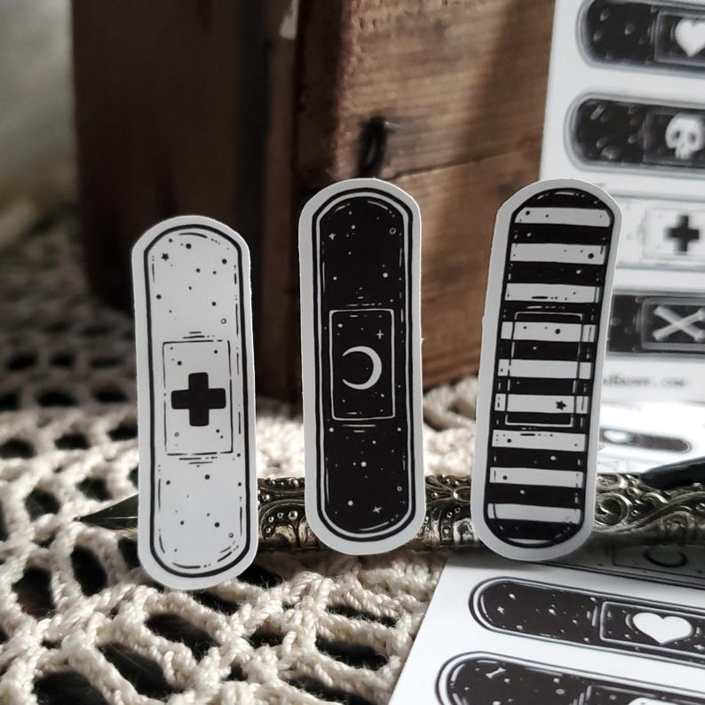May include: Three black and white band-aid stickers with different designs. The first sticker has a white background with a black cross and black dots. The second sticker has a black background with a white crescent moon and white stars. The third sticker has a black and white striped background with a white star.