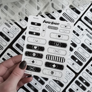 May include: A sheet of black and white paper bandages with various designs, including skulls, hearts, crosses, and moons. The sheet is titled "PAPER HAUNT: BANDAGES" and includes the text "By STAG".