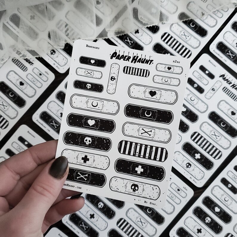 May include: A sheet of black and white paper bandages with various designs, including skulls, hearts, crosses, and moons. The sheet is titled "PAPER HAUNT: BANDAGES" and includes the text "By STAG".