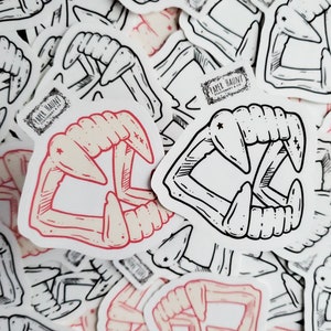 May include: A set of black and white stickers featuring a pair of fangs. The stickers are printed with the text "Paper Haunt".