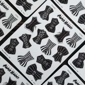 May include: A sheet of black and white stickers featuring various corset designs. The stickers are printed on white paper and have a glossy finish. The text "Paper Haunt" and "Corsets" is printed on the sheet.