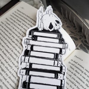 May include: Black and white sticker of a skull with candles on top of a stack of books. The sticker is on a white background.