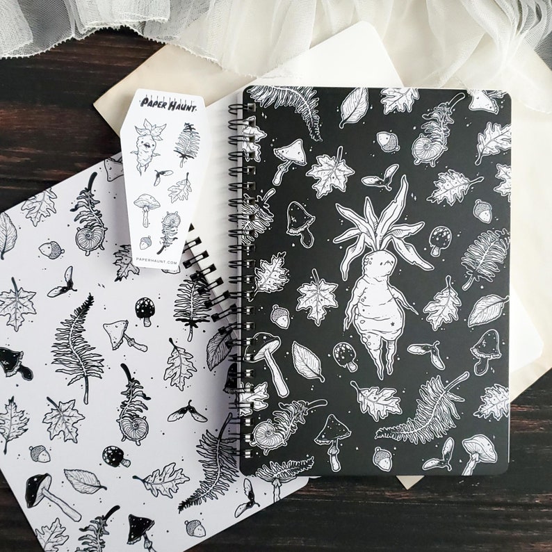 May include: A black and white spiral bound notebook with a whimsical illustration of a forest creature with leaves and mushrooms. The notebook cover features the text "PAPER HAUNT".