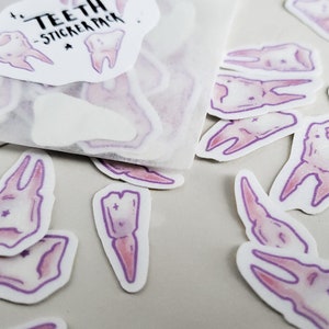 May include: A sheet of stickers featuring various tooth designs. The stickers are white with a purple outline and a pink fill. The sheet has the text "TEETH - STICKER PACK" printed on it.