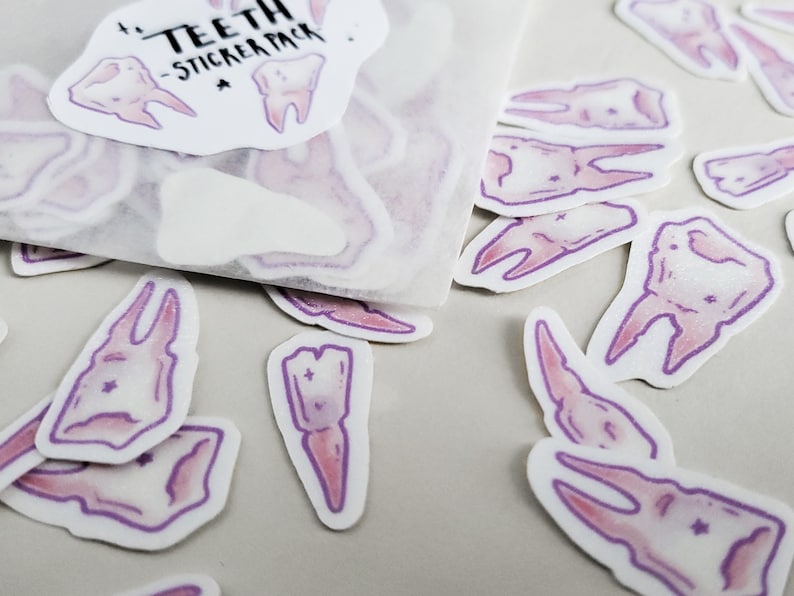 May include: A sheet of stickers featuring various tooth designs. The stickers are white with a purple outline and a pink fill. The sheet has the text "TEETH - STICKER PACK" printed on it.