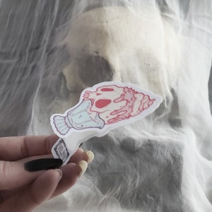 May include: A sticker featuring a stylized skull with pink and red accents, held against a blurred background. The skull is depicted with a decorative element, possibly a vase or container, in light blue. The sticker has a white border.