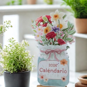 Puede incluir: Calendario floral de 2026 con diseño de tarro. El calendario presenta un ramo de flores coloridas y la palabra "Calendario". Se muestra sobre un pequeño caballete de madera, con plantas en macetas en el fondo.
