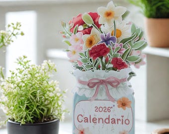 May include: A floral-themed 2026 calendar in a jar-shaped design. The calendar features a bouquet of colorful flowers and the word "Calendario". It is displayed on a small wooden easel, with potted plants in the background.