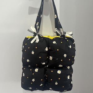 May include: A black and white floral tote bag with yellow trim and white ribbon bows. The bag has a quilted design and is made of fabric.