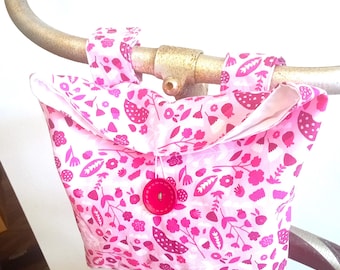 May include: A light pink fabric pouch featuring a dark pink floral and botanical pattern. The pouch has two fabric loops at the top for attachment and is secured in the centre with a single, round, dark pink button closure.