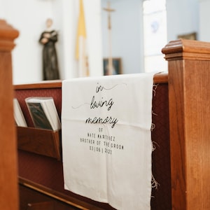 May include: White fabric banner with black text that reads "in loving memory of Nate Martinez, brother of the groom, 03|06|2022". The banner is hanging on a wooden pew in a church.