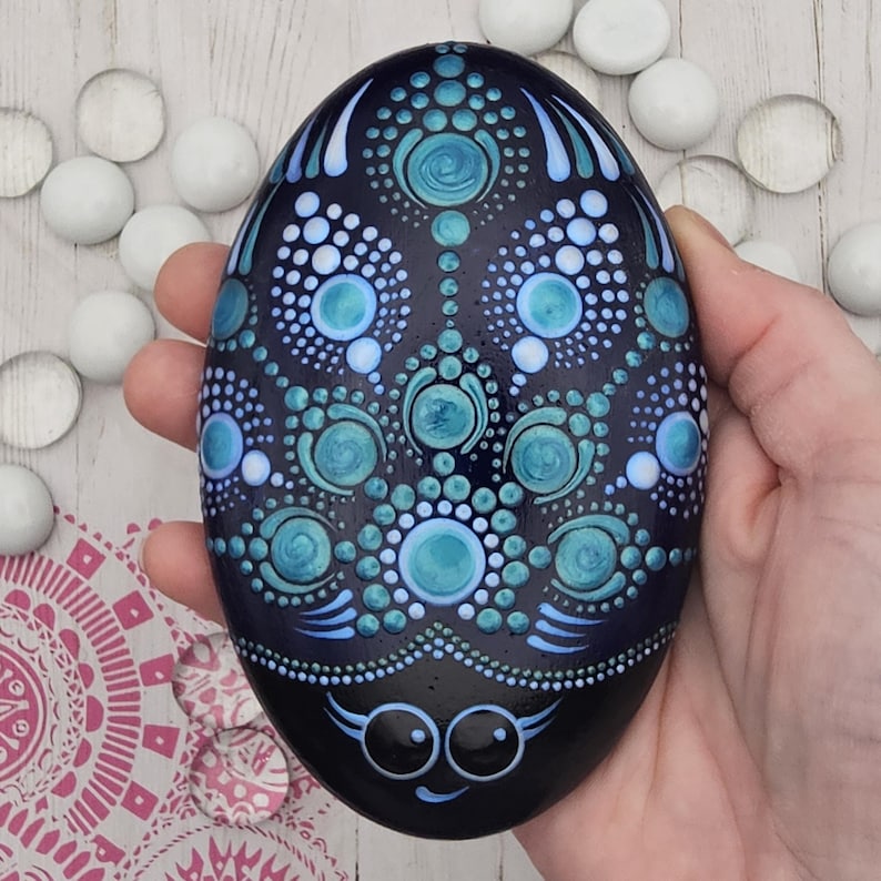 May include: A hand-painted, oval-shaped stone with a dark blue base, decorated with turquoise and light blue dot patterns. The design includes circular and teardrop shapes. The bottom of the stone has a smiling face with large eyes.