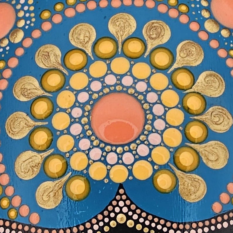 May include: Close-up of a hand-painted mandala design. The artwork features a central coral-colored circle surrounded by yellow and gold-colored circles and teardrop shapes. The background is a vibrant blue with smaller pink and white dots.