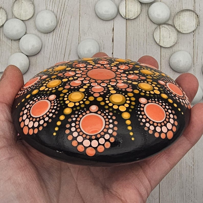 May include: A hand-painted, black stone featuring a detailed mandala design. The mandala incorporates orange, yellow, and peach-colored dots arranged in concentric circles. The stone is smooth and oval-shaped, showcasing a unique piece of art.
