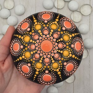 May include: A hand-painted, round black stone featuring a mandala design. The mandala is composed of orange, yellow, and peach-colored dots arranged in concentric circles. The stone is surrounded by white glass pebbles.