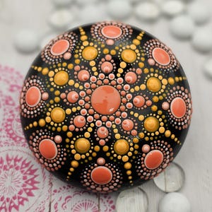May include: A hand-painted, round decorative stone with a black base. The stone features a mandala design in shades of orange, peach, and yellow. The design is composed of dots and circles, creating a symmetrical pattern. The stone is a unique piece of art.