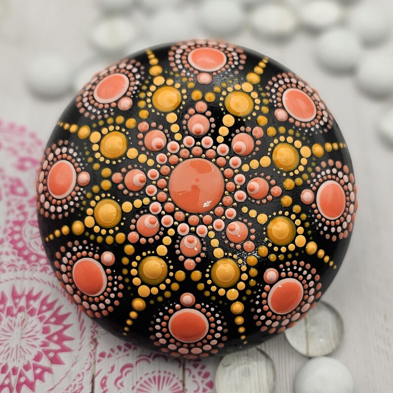 May include: A hand-painted, round decorative stone with a black base. The stone features a mandala design in shades of orange, peach, and yellow. The design is composed of dots and circles, creating a symmetrical pattern. The stone is a unique piece of art.