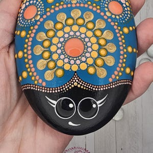 May include: Hand-painted decorative stone with a blue, orange, and gold mandala design. The bottom half is black with a smiling face. The stone is held in a hand, showcasing the intricate dot art.