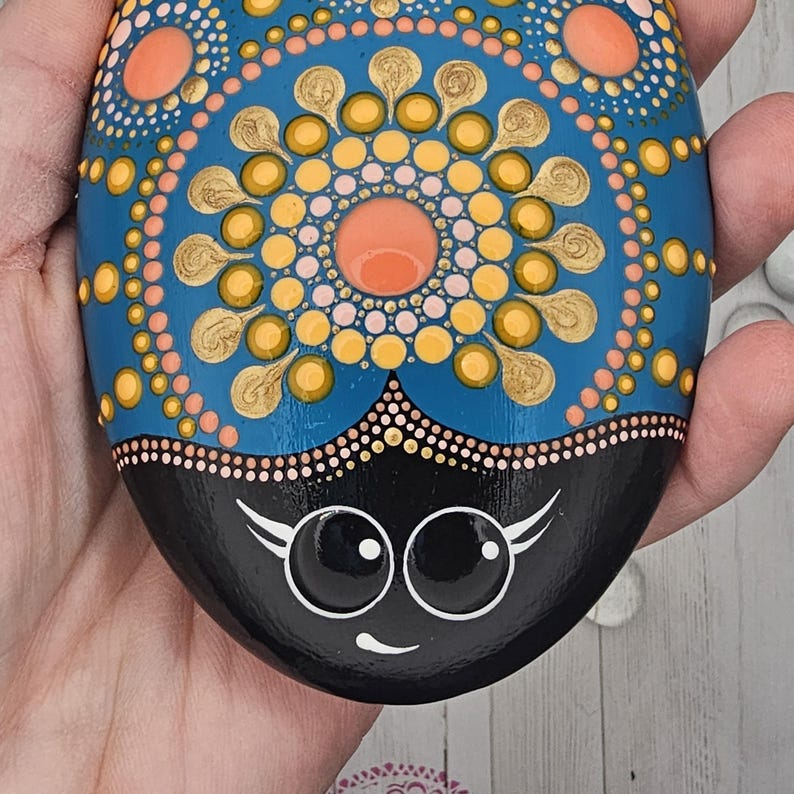 May include: Hand-painted decorative stone with a blue, orange, and gold mandala design. The bottom half is black with a smiling face. The stone is held in a hand, showcasing the intricate dot art.