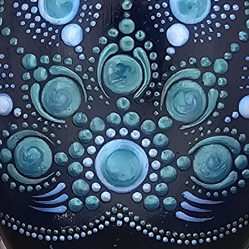 May include: Close-up of a dark blue ceramic vase with a hand-painted design. The design features teal and white dots and swirls arranged in a symmetrical pattern. The vase has a glossy finish.