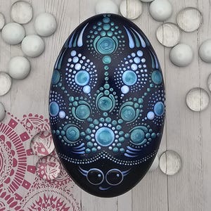 May include: A hand-painted, oval decorative stone. The dark blue stone features a detailed pattern of teal and light blue dots and circles. The bottom of the stone has a smiling face with eyelashes. White and clear glass beads surround the stone.
