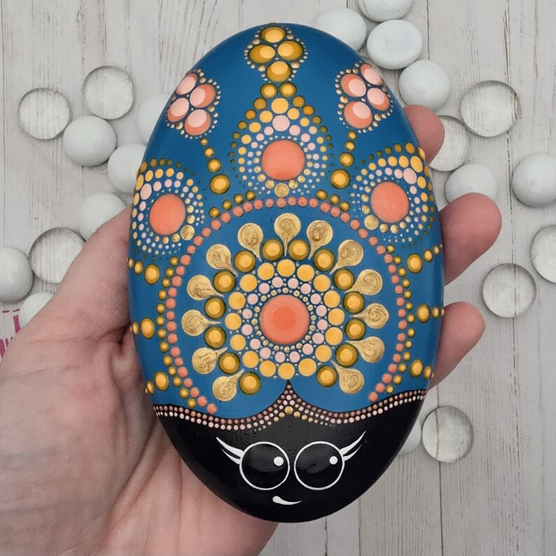 May include: A hand-painted, oval-shaped rock with a blue background and a detailed mandala design in shades of orange, yellow, and pink. The bottom is black with a cartoon ladybug face. Decorative stone art.