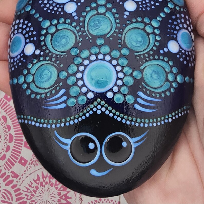 May include: A hand-painted, oval stone with a detailed dot art design. The stone is navy blue with turquoise and white accents. The design includes circular patterns and a stylized face at the bottom, creating a whimsical and decorative piece.