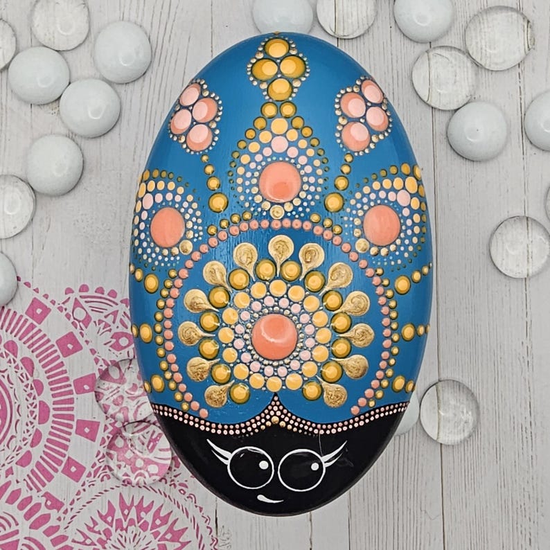 May include: A hand-painted, oval-shaped stone with a blue background and intricate dot patterns in gold, peach, and white. The bottom is black with a whimsical face. Decorative art piece.