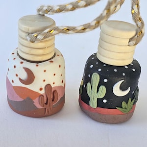 May include: Two small, decorative air freshener bottles with wooden tops and hanging cords. One bottle features a desert landscape with a crescent moon, the other a night sky with a cactus and moon. The bottles are hand-painted.