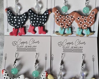 May include: Four pairs of dangle earrings on Copper Clover Clay Jewelry display cards. Each pair features two clay chickens with polka dots, wearing colorful boots and accented with a single clear or light green bead on silver-toned metal hooks.
