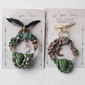 May include: Two circular pendants on display cards labeled Copper Clover Clay Jewelry. Each features a sculpted green snake coiled around a gold hoop adorned with brown branches, green leaves, and small mushrooms. One includes a clear crystal, the other a gold crescent moon charm.
