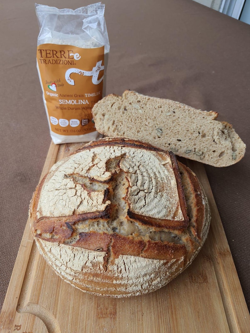May include: A loaf of sourdough bread with a crispy crust and a golden brown color. The bread is sitting on a wooden cutting board. A bag of organic ancient grain timilia semolina whole durum wheat flour is in the background.
